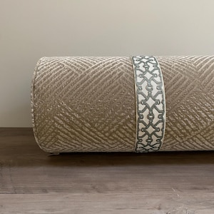 May include: A cylindrical decorative pillow with a textured beige fabric. A band of white fabric with a gray geometric pattern encircles the pillow. The pillow is resting on a wooden surface.