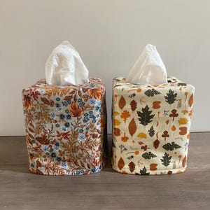 May include: Two square tissue box covers. One features a floral pattern with blue, orange, and brown flowers on a beige background. The other has a cream background with autumn leaves and mushroom designs.