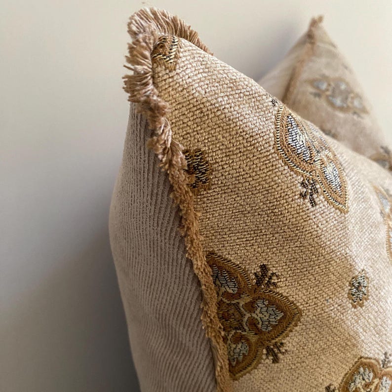 May include: Close-up of a decorative pillow with a beige background. The pillow features a patterned design in gold, brown, and blue. The edges are trimmed with a fringe in a matching gold color. The pillow has a soft, textured appearance.