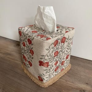 May include: A square tissue box cover with a botanical pattern of red pomegranates and gray leaves on a beige background. The cover has a fringed trim at the base and a white tissue sticking out of the top.