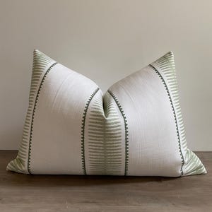 May include: A decorative throw pillow with a white background and a vertical stripe pattern in a muted green color. The pillow has a textured appearance and is designed for home decor.