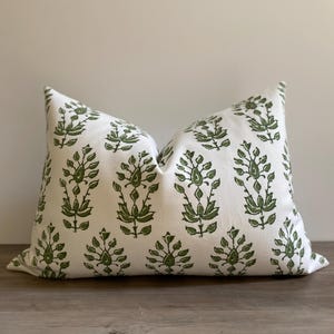 May include: A rectangular decorative pillow with a white background and a repeating pattern of green floral designs. The pillow is placed on a wooden surface and has a soft, inviting appearance. The pillow is designed for home decor.