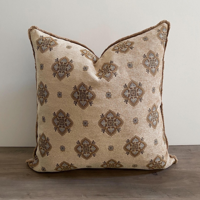 May include: Square decorative pillow with a beige background and a repeating pattern of brown and gray floral designs. The pillow has a brown fringe trim around the edges, adding a textured detail. The pillow is a home decor item.