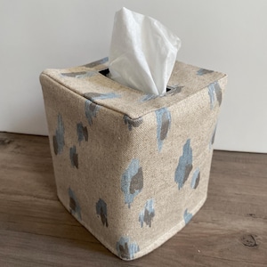 May include: A square tissue box cover with a beige fabric featuring blue and brown abstract brushstroke patterns. White tissues are pulled from the top opening. The box is sitting on a wooden surface.
