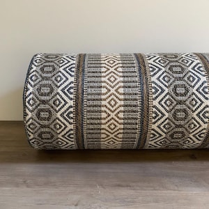 May include: A cylindrical pillow with a woven fabric cover. The pillow features a repeating geometric pattern in shades of brown, gray, and white. The design includes diamond and rectangular shapes, creating a textured appearance. The pillow is designed for comfort and decorative use.