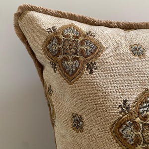 May include: Close-up of a beige decorative pillow with a repeating geometric pattern in shades of brown and gold. The pillow has a textured fabric and is trimmed with a fringe in a matching brown color. The pillow is designed for home decor.