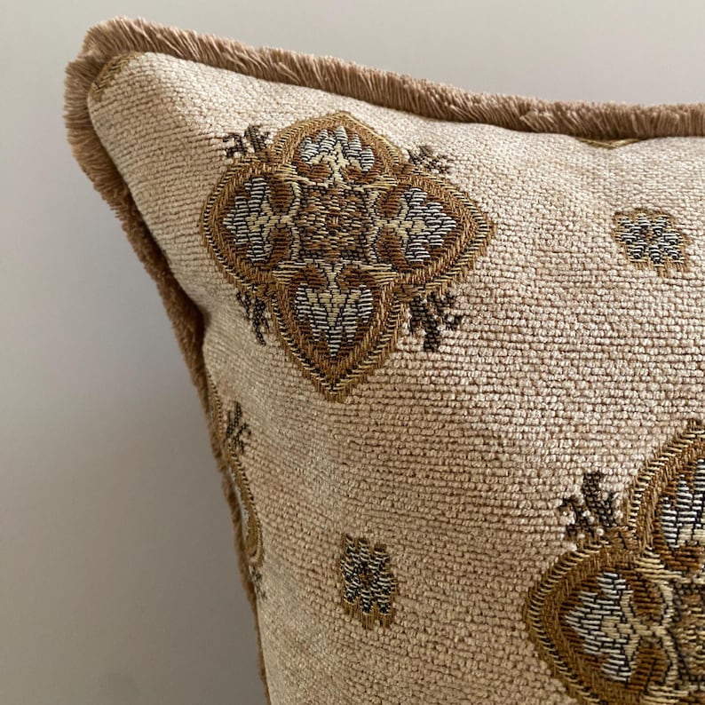 May include: Close-up of a beige decorative pillow with a repeating geometric pattern in shades of brown and gold. The pillow has a textured fabric and is trimmed with a fringe in a matching brown color. The pillow is designed for home decor.