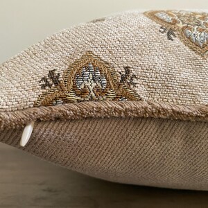 May include: Close-up of a decorative pillow with a beige woven fabric. The pillow features intricate embroidered patterns in shades of brown, gold, and blue. The edges are trimmed with a fringed border, and a zipper is visible on the side.