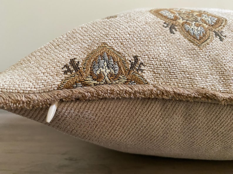 May include: Close-up of a decorative pillow with a beige woven fabric. The pillow features intricate embroidered patterns in shades of brown, gold, and blue. The edges are trimmed with a fringed border, and a zipper is visible on the side.