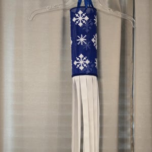 May include: A cylindrical, blue and white decorative hanging ornament. The ornament features a pattern of white snowflakes on a blue background. White fringe hangs from the bottom. The ornament is suspended from a clear plastic hanger with a blue ribbon.
