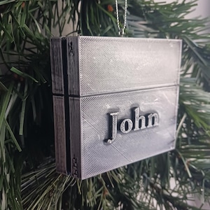 May include: A silver-colored, 3D printed Christmas ornament shaped like a video game console with the name "John" printed on the front.