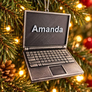 Pode incluir: Um enfeite de árvore de Natal em formato de computador portátil com acabamento prateado metálico e tela preta. O nome Amanda é exibido em letras prateadas em relevo na tela. O enfeite apresenta um design detalhado de teclado e trackpad.
