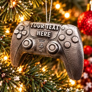 May include: A silver-toned, metallic gaming controller ornament hanging from a thin chain. The controller features a textured surface with the embossed text "YOUR TEXT HERE" and a small PlayStation logo centred between the two analogue sticks.