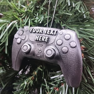 May include: A silver-coloured PlayStation 5 controller ornament with the text "YOUR TEXT HERE" on the front. The ornament is hanging from a Christmas tree.