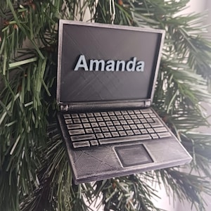 以下が含まれることがあります： 黒いラップトップのオーナメントで、銀色のキーボードと画面に「Amanda」という名前が印刷されています。オーナメントはクリスマスツリーにぶら下がっています。
