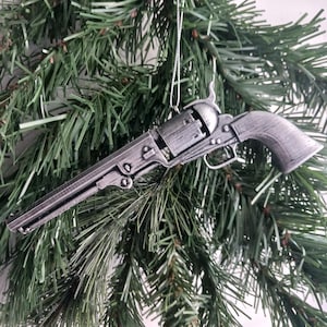 Pode incluir: Um ornamento de revólver prateado pendurado em uma árvore de Natal. O ornamento é detalhado e realista, com um acabamento texturizado.