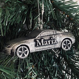 May include: Silver car ornament with the name "Maria" in black lettering. The ornament is hanging from a Christmas tree. The car has detailed wheels and a textured finish, giving it a realistic appearance.