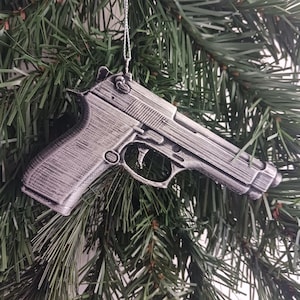 May include: A silver-colored pistol-shaped Christmas tree ornament. The ornament is detailed with realistic features and hangs from a silver string against a backdrop of green pine needles.