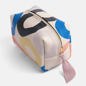 Könnte beinhalten: Eine beige Reißverschlusstasche mit einem farbenfrohen abstrakten Design mit blauen, rosa, gelben und schwarzen Kreisen. Die Tasche hat einen rosa Schleifen-Reißverschluss.