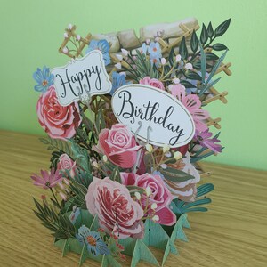 Peut inclure: Une carte pop-up 3D sur le th&egrave;me floral. La carte pr&eacute;sente des roses roses, des fleurs bleues et des feuilles vertes. La carte est d&eacute;cor&eacute;e de deux pancartes qui disent "Joyeux anniversaire".