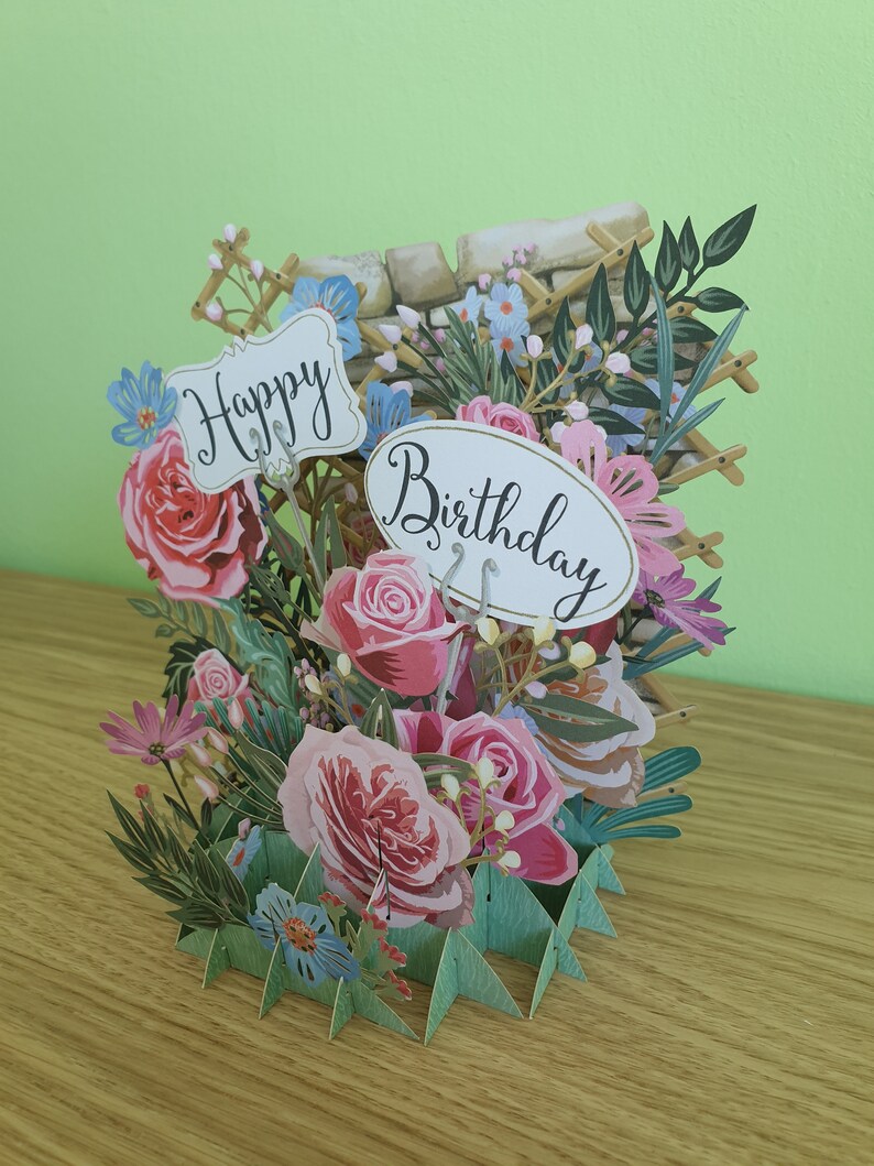 Peut inclure: Une carte pop-up 3D sur le th&egrave;me floral. La carte pr&eacute;sente des roses roses, des fleurs bleues et des feuilles vertes. La carte est d&eacute;cor&eacute;e de deux pancartes qui disent "Joyeux anniversaire".