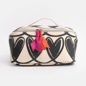 Könnte beinhalten: Cremefarbene Kosmetiktasche mit schwarzem Herzmuster. Die Tasche hat einen oberen Griff, einen Reißverschluss und eine dekorative Quaste mit rosa, orangefarbenen und fuchsiafarbenen Bändern. Die Tasche ist rechteckig und hat einen neutralen Hintergrund.
