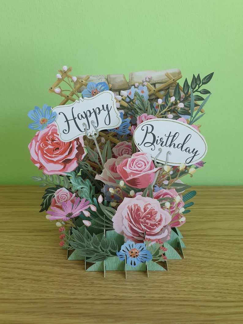 Peut inclure: Une carte pop-up 3D sur le th&egrave;me floral. La carte pr&eacute;sente un bouquet de roses roses et blanches, de fleurs bleues et de feuilles vertes. La carte comporte une pancarte blanche qui dit "Happy" et une pancarte ovale blanche qui dit "Birthday".
