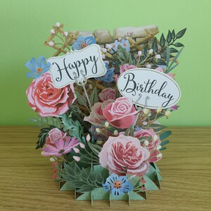 Peut inclure: Une carte pop-up 3D sur le th&egrave;me floral. La carte pr&eacute;sente des roses roses, des fleurs bleues et des feuilles vertes. La carte est ouverte et les mots "Happy Birthday" sont visibles.