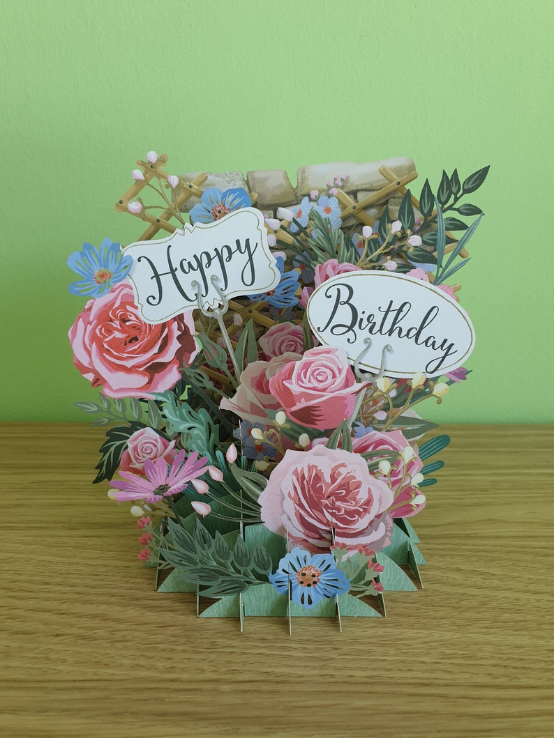 Peut inclure: Une carte pop-up 3D sur le th&egrave;me floral. La carte pr&eacute;sente des roses roses, des fleurs bleues et des feuilles vertes. La carte est ouverte et les mots "Happy Birthday" sont visibles.