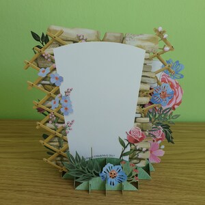 Peut inclure: Une carte pop-up repr&eacute;sentant un mur en pierre avec un treillis en bois et des fleurs dans des tons de rose, bleu et vert. La carte est vierge &agrave; l'int&eacute;rieur pour &eacute;crire un message.
