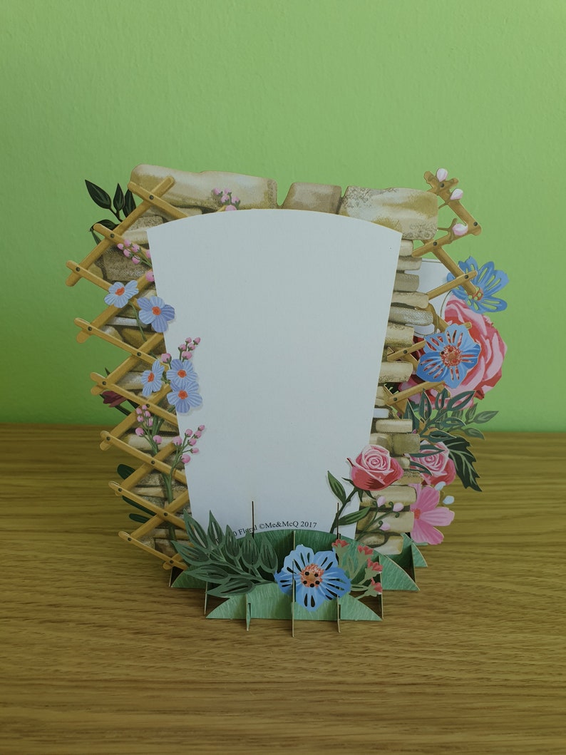 Peut inclure: Une carte pop-up repr&eacute;sentant un mur en pierre avec un treillis en bois et des fleurs dans des tons de rose, bleu et vert. La carte est vierge &agrave; l'int&eacute;rieur pour &eacute;crire un message.
