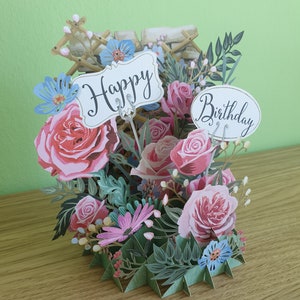 Peut inclure: Une carte 3D pop-up avec un bouquet de fleurs. La carte pr&eacute;sente des roses roses, des fleurs bleues et des feuilles vertes. La carte a une pancarte blanche qui dit "Joyeux anniversaire".
