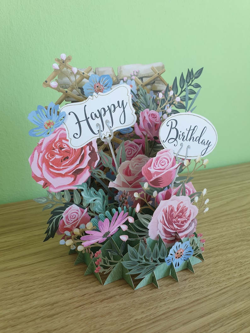 Peut inclure: Une carte 3D pop-up avec un bouquet de fleurs. La carte pr&eacute;sente des roses roses, des fleurs bleues et des feuilles vertes. La carte a une pancarte blanche qui dit "Joyeux anniversaire".