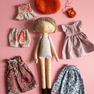 Könnte beinhalten: Eine handgemachte Puppe mit braunen Haaren und einem gestreiften Shirt, zusammen mit einer Vielzahl von Kleidungsstücken, darunter Blumenkleider, ein Rock, Shorts und ein Hut.