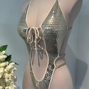 Op de afbeelding: Een eendelig bodysuit met een reflecterend zilverkleurig zeshoekig patroon, een diepe V-hals met een beige vetersluiting, dunne verstelbare beige bandjes en hoog uitgesneden beenopeningen, getoond op een paspop.