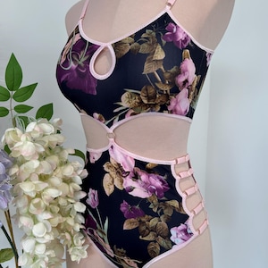 Könnte beinhalten: Ein zweiteiliges Dessous-Set mit schwarzem Hintergrund und einem floralen Muster in Rosa- und Lilatönen. Das Oberteil hat ein Schlüssellochdetail, und die Hose hat seitliche Ausschnitte mit rosa Besatz. Das Set wird auf einer Büste präsentiert.