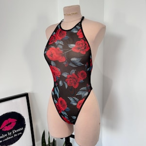 Puede incluir: Un body negro con cuello halter y un estampado floral de rosas rojas. La tela transparente presenta grandes rosas rojas con hojas verdes. El body se exhibe en un maniquí. El texto "Desire by Donna" es visible en el fondo.
