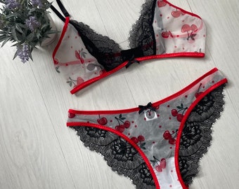 May include: A lingerie set featuring a sheer bralette and matching panties. The set is white with a cherry pattern, black lace trim, and red accents. Both pieces have small black bows.