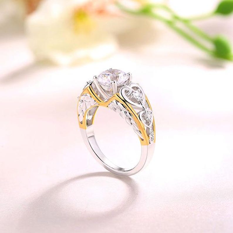 May include: A silver and gold ring with a large, clear gemstone at the center. The ring features intricate filigree detailing and smaller accent stones. The band is silver, with gold accents along the edges and around the main stone.