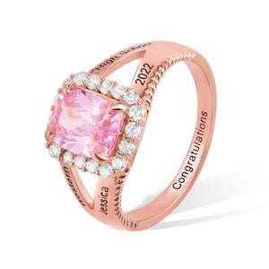 May include: Rose gold ring with a pink gemstone surrounded by smaller clear gemstones. The ring has the inscription "Jessica High School 2022" on one side and "Congratulations" on the other.