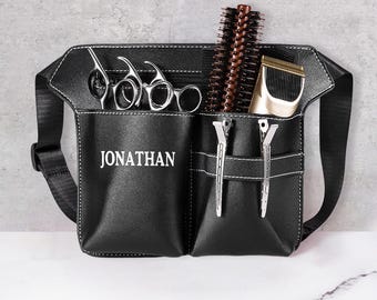 Puede incluir: Cinturón de herramientas de peluquería de cuero negro con costuras blancas y el nombre "JONATHAN" impreso en la parte delantera. El cinturón contiene tijeras, peines, pinzas para el pelo y una recortadora. El cinturón tiene una correa ajustable.
