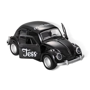 May include: Black toy car model of a classic Volkswagen Beetle with a white inscription that says "Tess" and the date "09-23-2021" on the roof. The car has a door open and the headlights are visible.