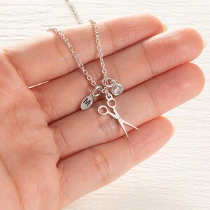 May include: A silver-colored necklace featuring a small pair of scissors, a round charm with the letter "M", and a clear crystal pendant. The necklace is displayed against a light background.