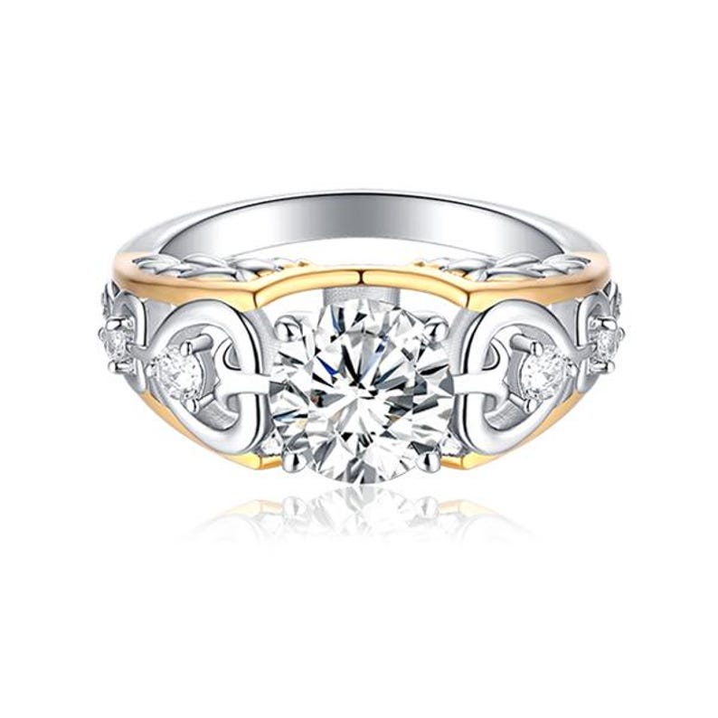 May include: A silver and gold ring with a large, clear, round-cut center stone. The ring features a decorative band with smaller accent stones and a gold-colored inner band. The design is intricate and elegant, suitable for special occasions.