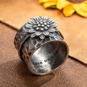 Peut inclure: Une bague en argent avec un motif de tournesol. La bague présente un tournesol détaillé sur le dessus et un motif géométrique sur le bandeau. L'intérieur de la bague est gravé des mots "Love from Nanny".