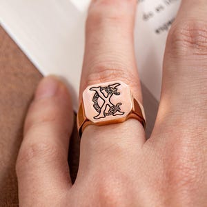 May include: A rose gold signet ring with a square face. The face of the ring features a black engraved design of the letter "X" with an ornate, stylized pattern. The ring is worn on a finger.