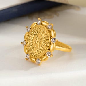 May include: A gold-toned ring featuring a central oval medallion with the text LOCK HAVEN UNIVERSITY and 2025, surrounding the letters NS framed by laurel leaves. The medallion is bordered by alternating smooth gold ovals and clear round stones.