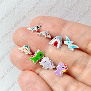 Puede incluir: Una colección de pequeños pendientes de botón con temática de dinosaurios. Los pendientes presentan varios diseños de dinosaurios, incluyendo un T-Rex, un Triceratops y un Pterodáctilo, en tonos verdes, morados y azules. Los pendientes son de metal.