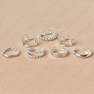 May include: A collection of silver-colored rings in various designs, including twisted, beaded, and openwork styles. The rings are displayed on a light brown surface, showcasing their different textures and shapes.
