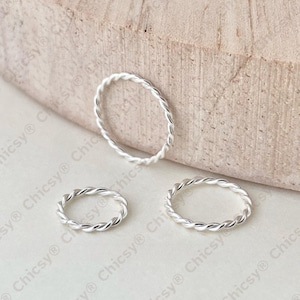 May include: Three silver-coloured twisted rings. One ring is standing upright, while the other two are lying flat. The rings are on a light-coloured surface with a wooden background.
